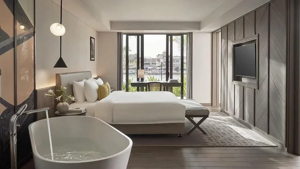 Junior Grand Suite tại Moire Hoi An, bồn tắm trắng đặt đầu phòng, cửa kính lớn nhìn ra sông Hoài và dãy nhà phố cổ