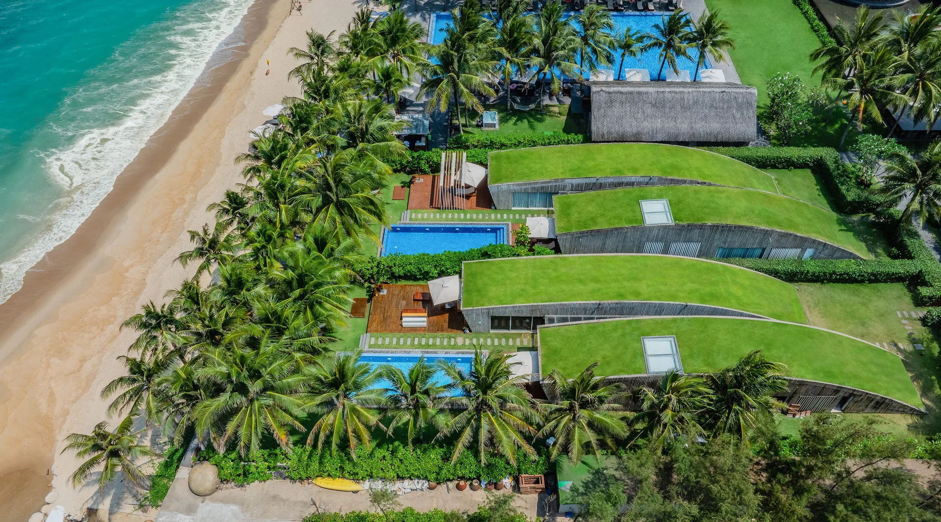 Naman Retreat nhìn từ trên cao: các villa mái cỏ xanh, hồ bơi riêng giữa rừng dừa, sát bãi cát và sóng biển Non Nước
