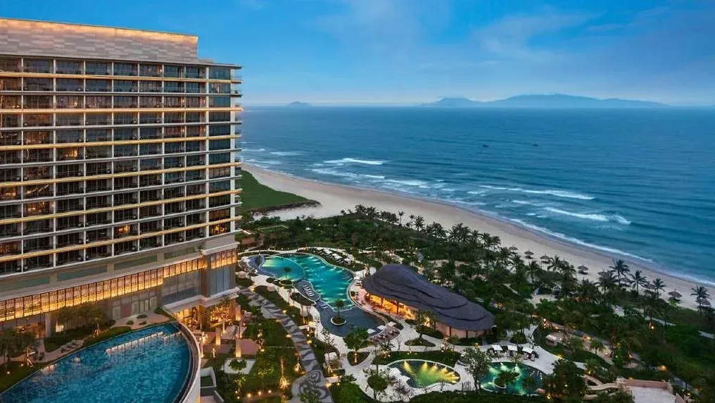 Toà tháp New World Hoiana Beach Resort lên đèn lúc chiều, bên dưới là hệ hồ bơi uốn lượn giữa vườn dừa và bãi biển dài