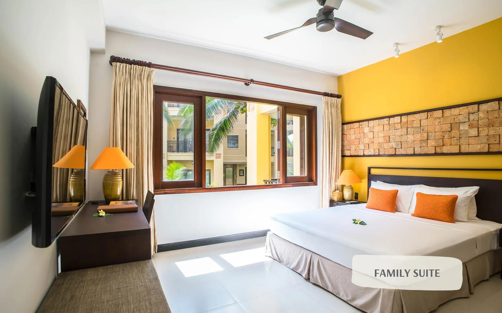 Phòng Family Suite tại Pandanus Resort với tường vàng, mảng ốp đá ong phía đầu giường, giường lớn và cửa sổ nhìn ra hàng dừa
