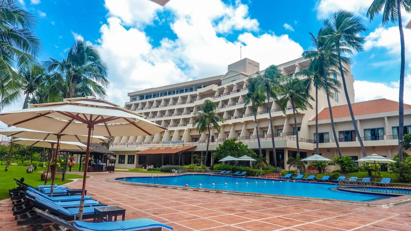 Khối nhà bậc thang của Phan Thiet Ocean Dunes Resort sau hồ bơi ngoài trời, hàng dù và ghế tắm nắng giữa vườn dừa