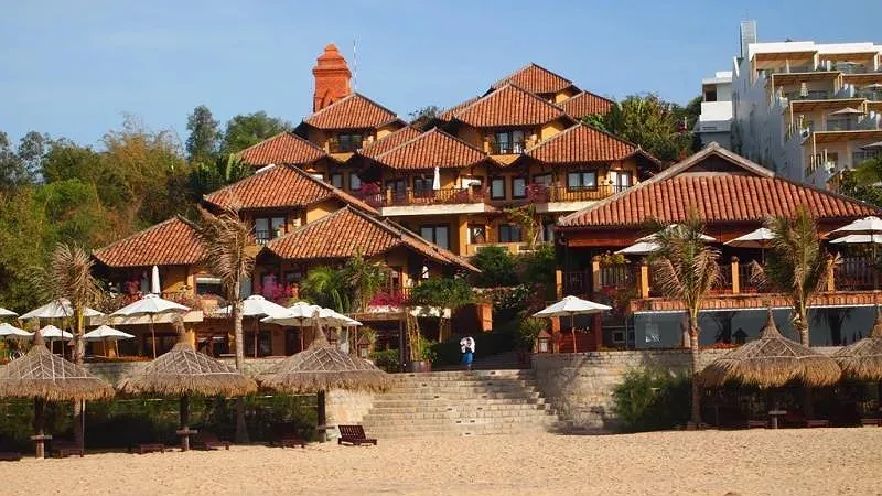 Các dãy nhà mái ngói đỏ kiểu làng Chăm của Poshanu Resort với tháp gạch phía sau, bậc đá dẫn xuống bãi cát có chòi lá
