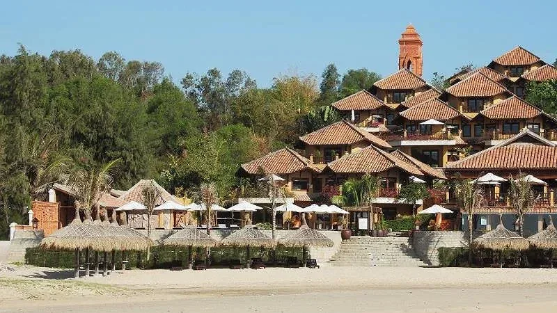 Dãy nhà mái ngói đỏ và tháp gạch kiểu Chăm của Poshanu Resort trên triền đồi, phía trước là chòi lá trên bãi cát