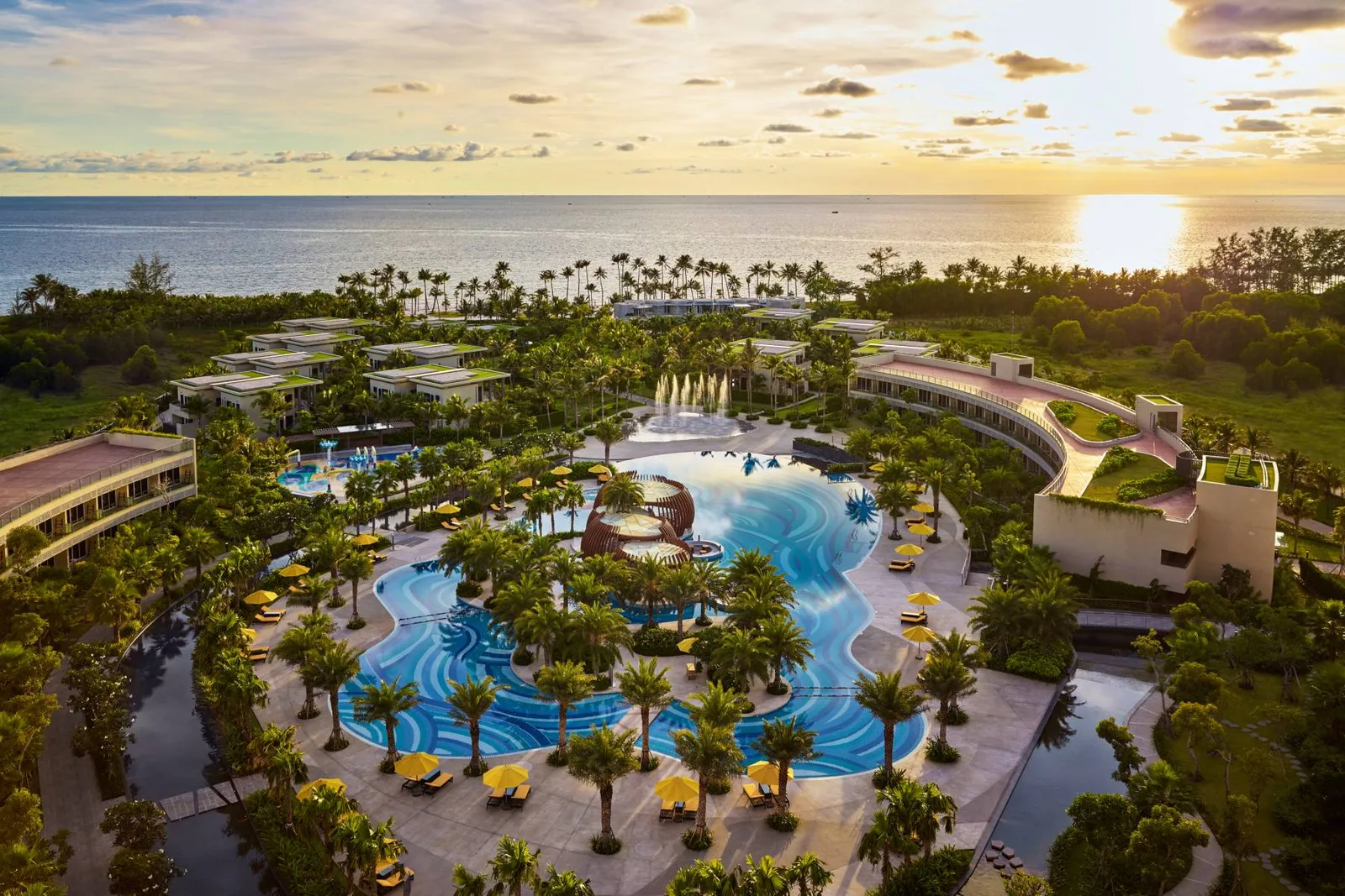 Pullman Phu Quoc Beach Resort nhìn từ trên cao lúc hoàng hôn: hồ bơi uốn lượn hoa văn sóng giữa hàng dừa và dù vàng, phía sau là biển