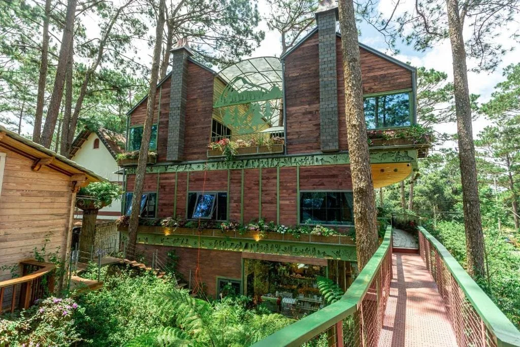 Nhà gỗ nhiều tầng của Rosie Garden Măng Đen với mái kính, bồn hoa trên ban công và cầu gỗ lan can xanh dẫn vào giữa rừng thông