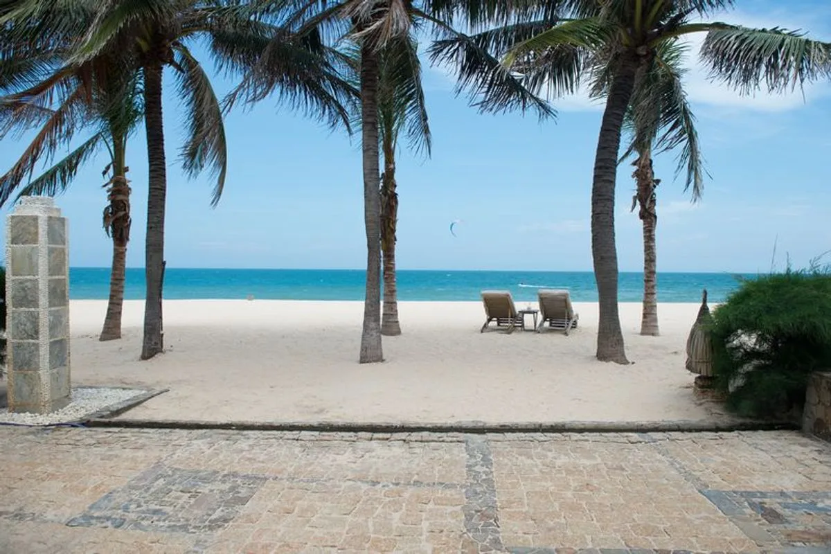 Bãi cát trắng trước Sailing Club Resort Mũi Né, hai ghế nằm dưới hàng dừa, xa xa có cánh diều lướt ván trên biển