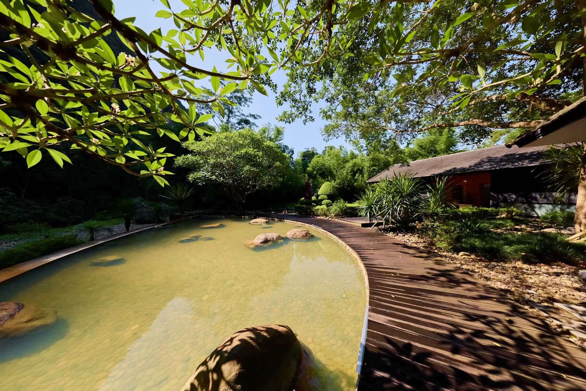 Bể khoáng ngoài trời đáy sỏi có đá tảng, lối đi gỗ cong và nhà gỗ mái ngói ở khu Onsen kiểu Nhật, Serena Resort Kim Bôi
