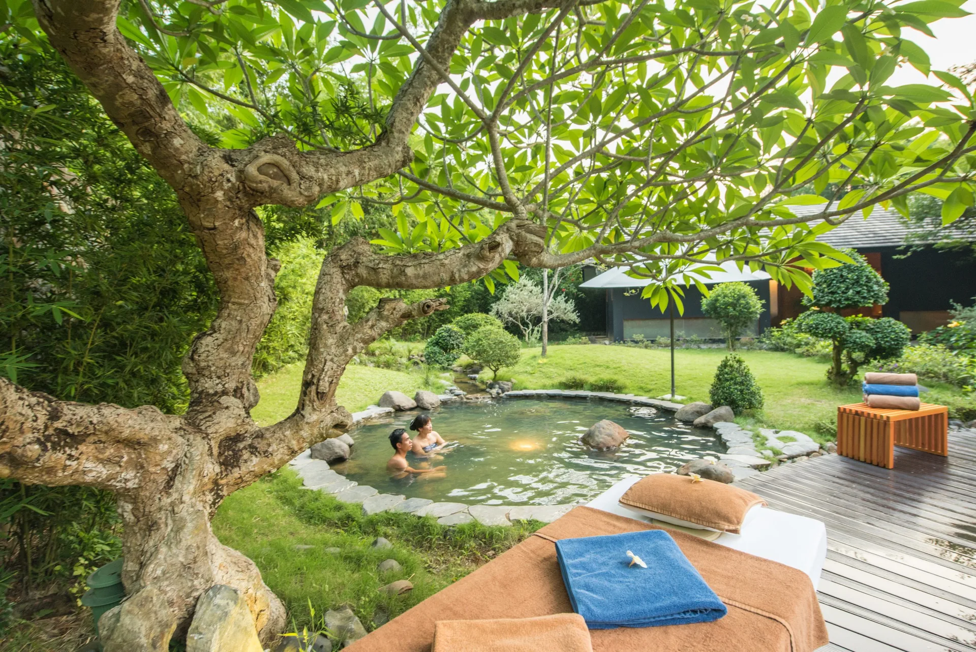 Hai khách ngâm trong bể khoáng ngoài trời viền đá dưới gốc cây đại tại khu Onsen kiểu Nhật của Serena Resort Kim Bôi