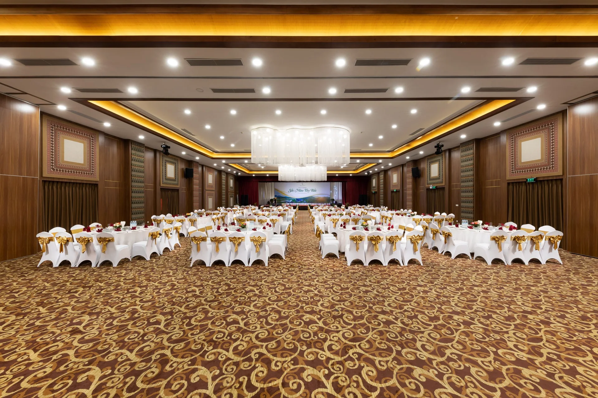 Phòng tiệc lớn tại Serena Resort Kim Bôi kê bàn tròn khăn trắng, ghế nơ vàng, đèn chùm pha lê và vách hoa văn thổ cẩm