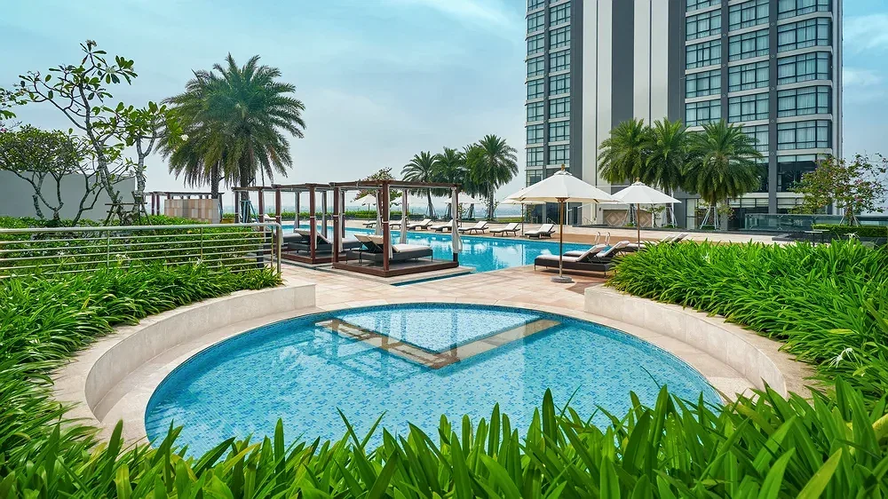 Hồ bơi ngoài trời tròn và hồ bơi dài giữa vườn cây, cabana và ô dù trắng, tòa tháp Sheraton Cần Thơ Hotel phía sau