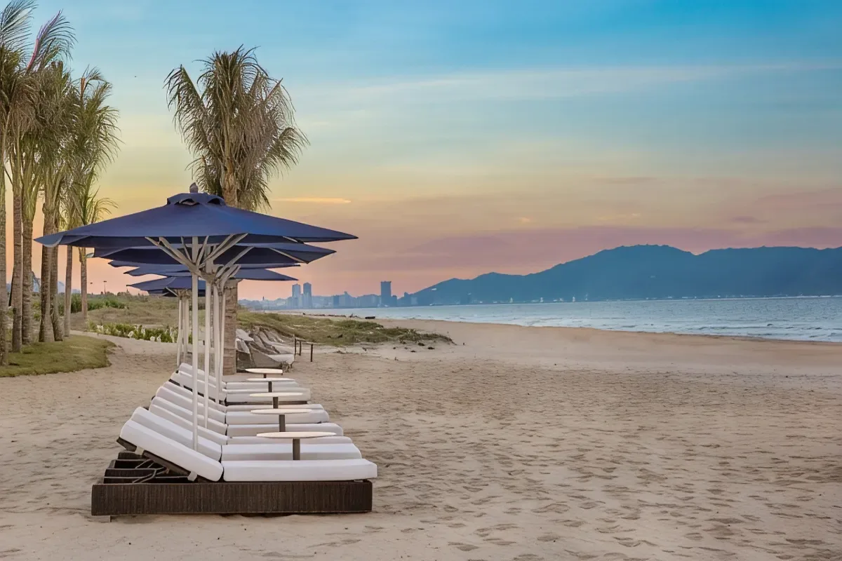Hàng ghế tắm nắng trắng và dù xanh trên bãi cát riêng của Sheraton Grand Danang lúc hoàng hôn, xa xa là bán đảo Sơn Trà