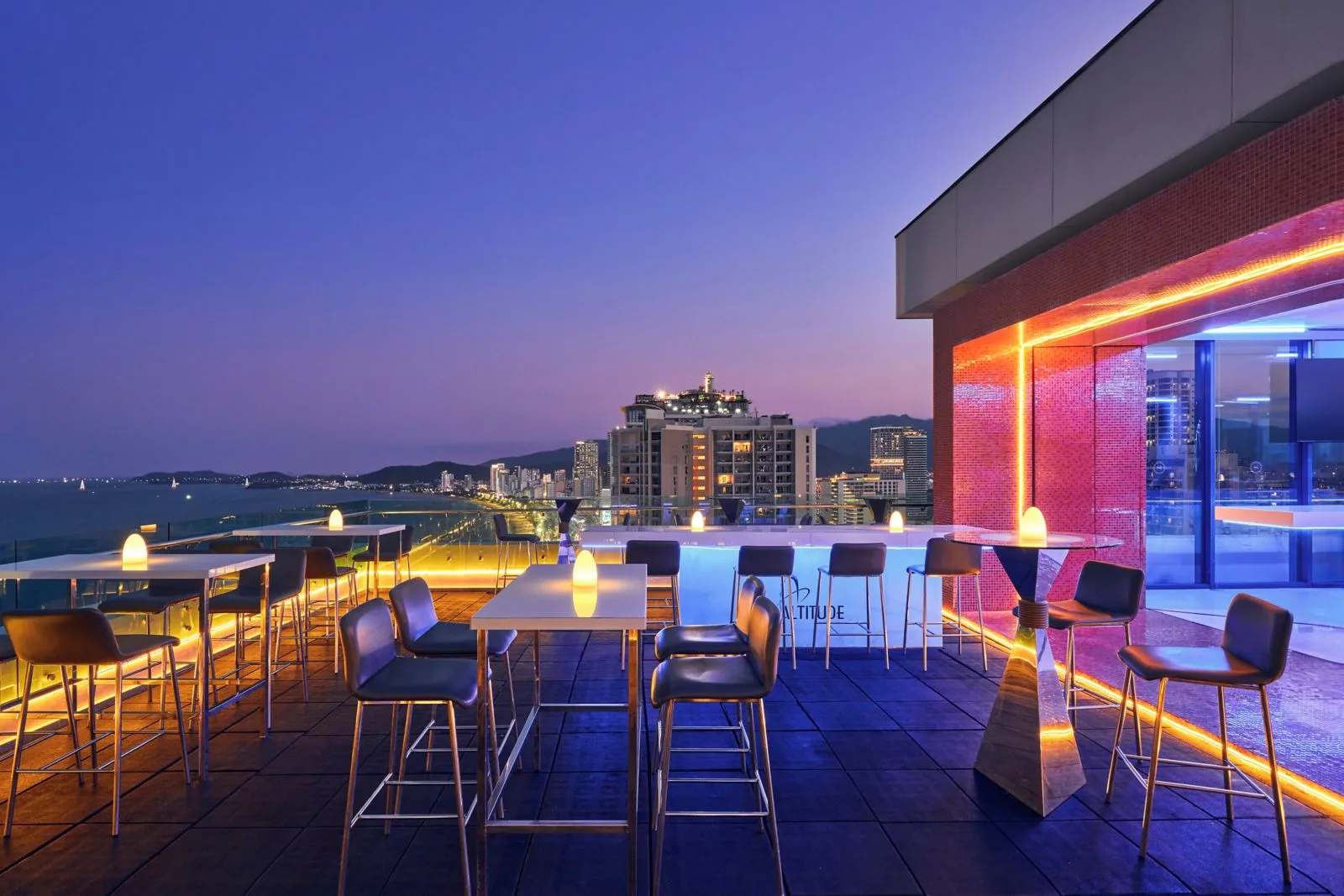 Altitude Rooftop Bar của Sheraton Nha Trang Hotel & Spa lúc chạng vạng, dãy ghế cao hướng ra thành phố và mặt vịnh lên đèn