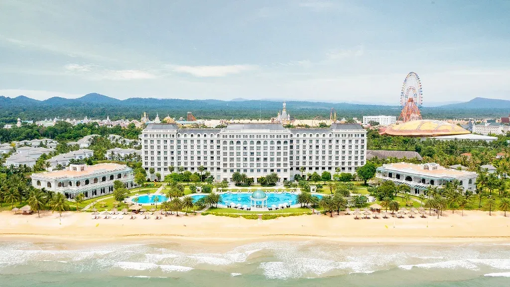 Sheraton Phu Quoc Long Beach Resort nhìn từ biển: tòa nhà chính màu trắng, hồ bơi có đài mái vòm ở giữa bãi cỏ, dãy ô trên bãi cát, xa xa là vòng quay và dãy núi