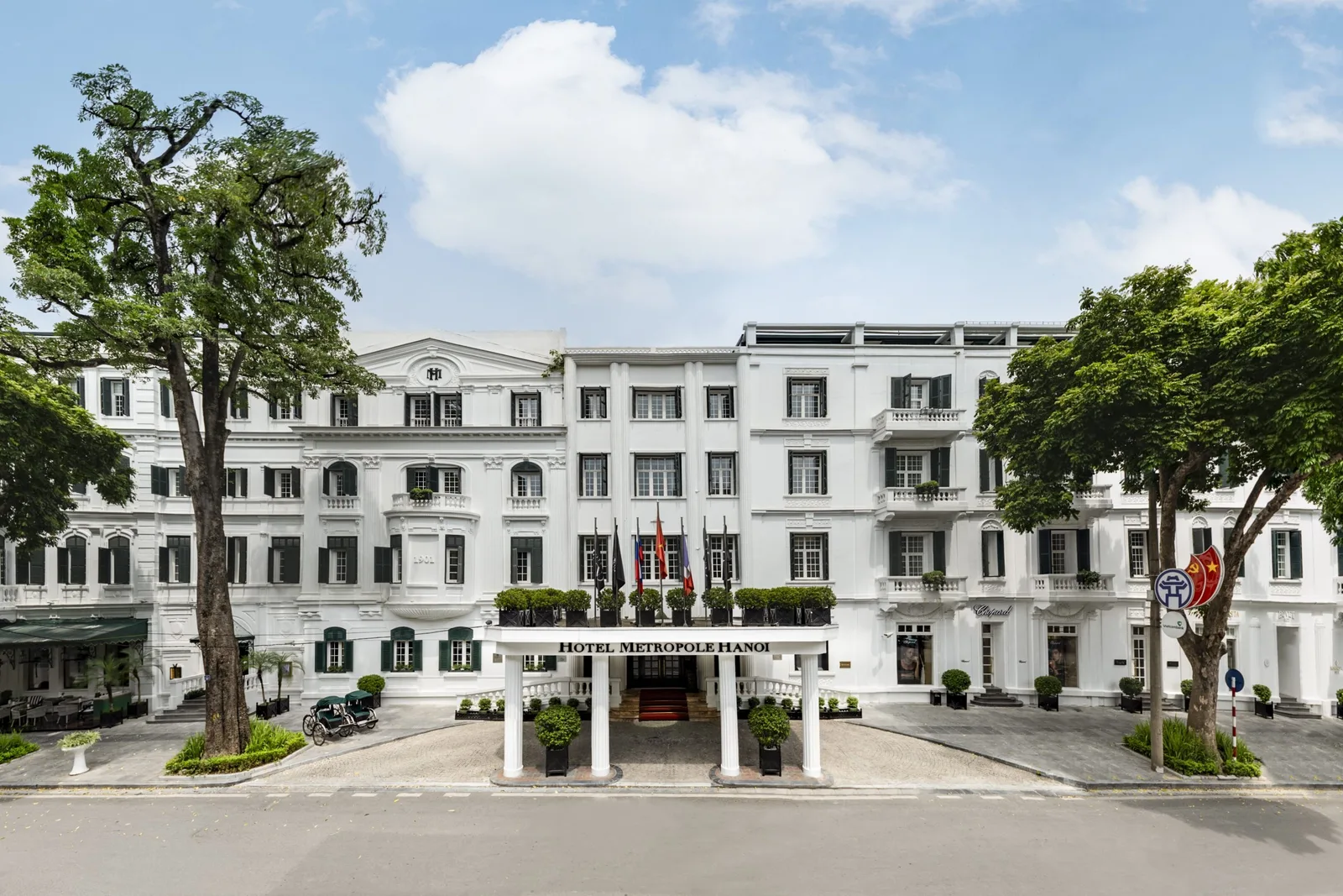 Mặt tiền trắng kiểu Pháp của Sofitel Legend Metropole Hanoi, cửa chớp xanh, hàng cờ và cây xanh trên phố Ngô Quyền