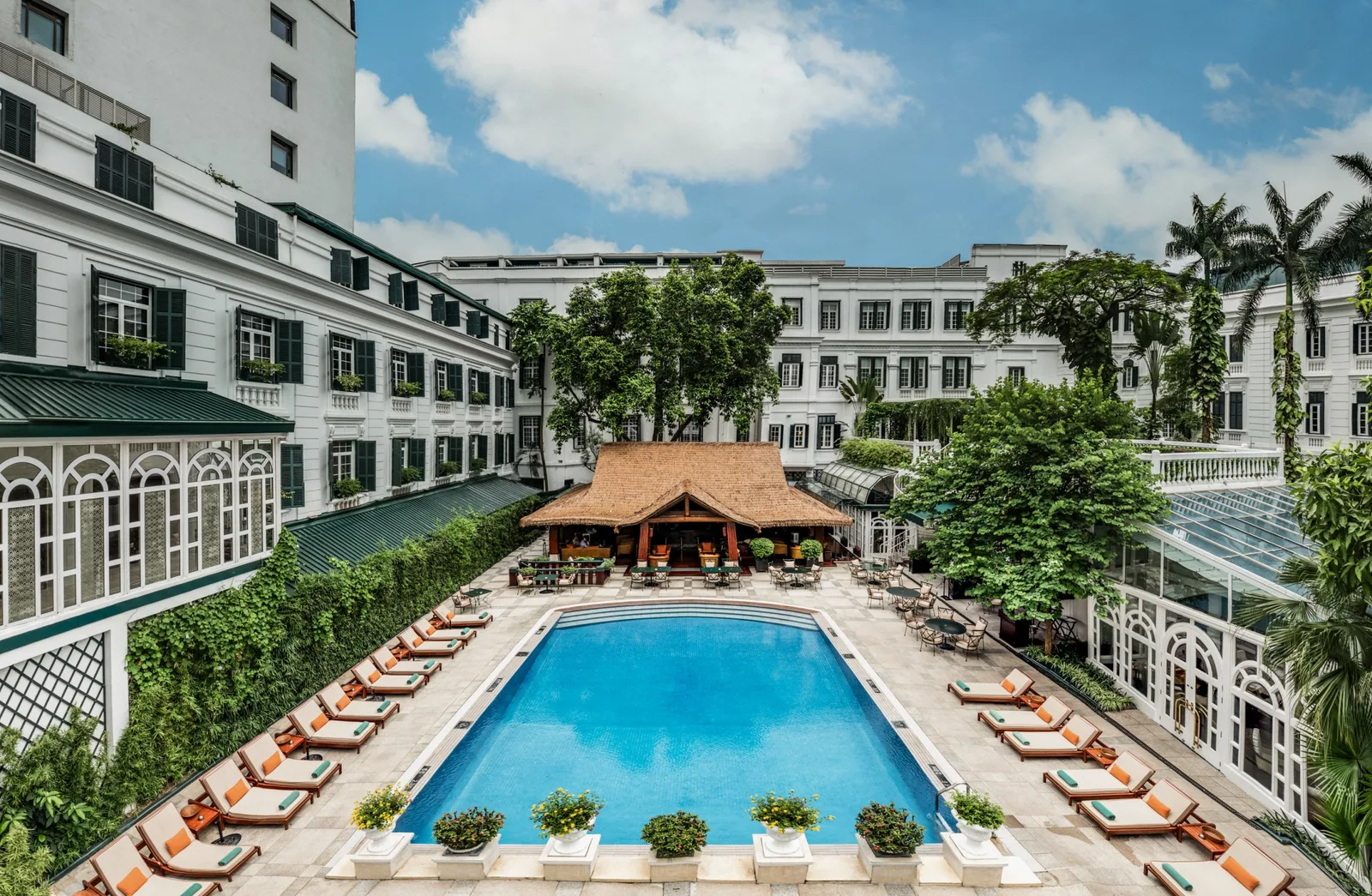 Sân trong Sofitel Legend Metropole Hanoi với bể bơi xanh, hàng ghế tắm nắng, quầy bar mái lá giữa các dãy nhà trắng cửa chớp xanh