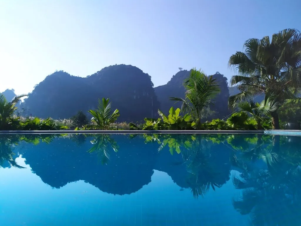 Bể bơi vô cực phản chiếu dãy núi đá vôi và hàng cọ dưới trời xanh tại Tam Coc La Montagne Resort & Spa