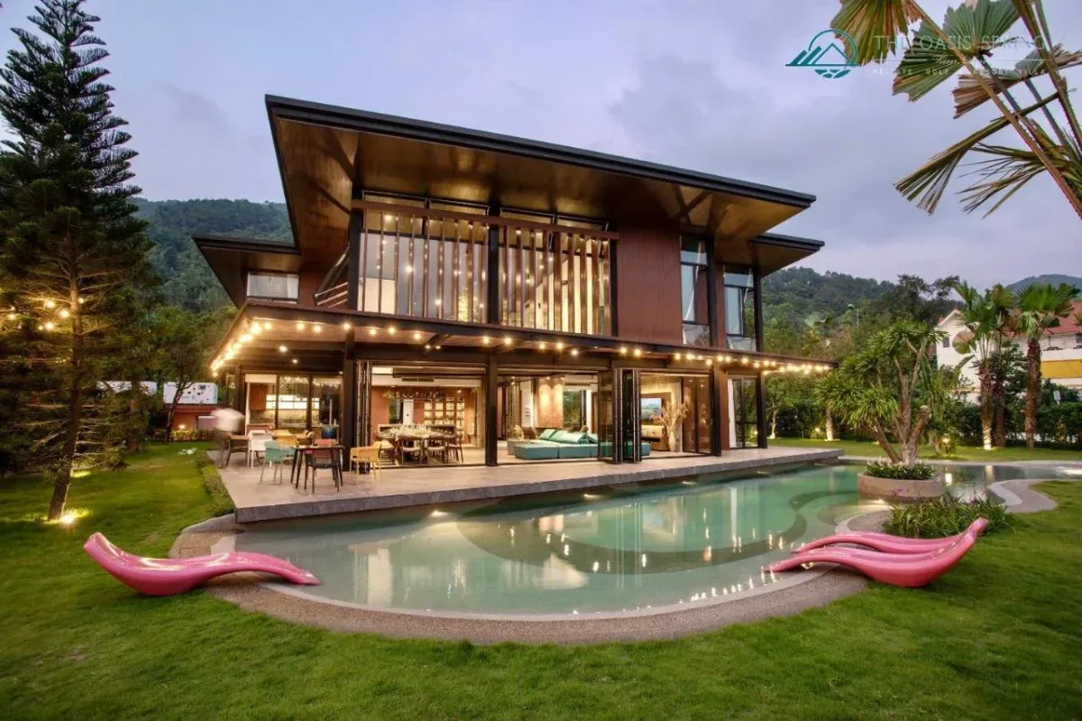 Villa hai tầng ốp gỗ của Tam Đảo Golf & Resort lúc chiều tối, hồ bơi riêng uốn lượn trên bãi cỏ, dây đèn và núi phía sau