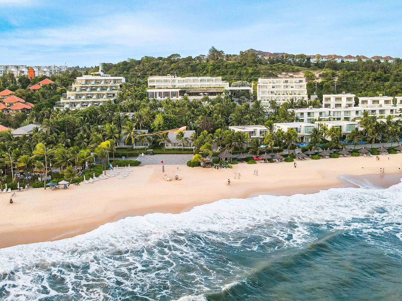 Toàn cảnh The Cliff Resort & Residences nhìn từ biển: dãy villa và khối nhà trắng dưới rặng dừa, bãi cát vàng có dù và ghế nằm, sóng vỗ phía trước