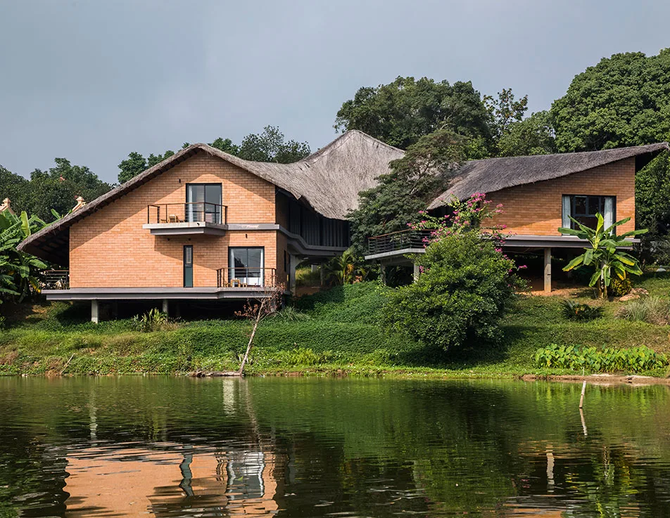 Hai căn villa tường gạch mộc, mái lá dày của Tomodachi Retreat - Muong Village nằm trên triền cỏ sát mặt hồ