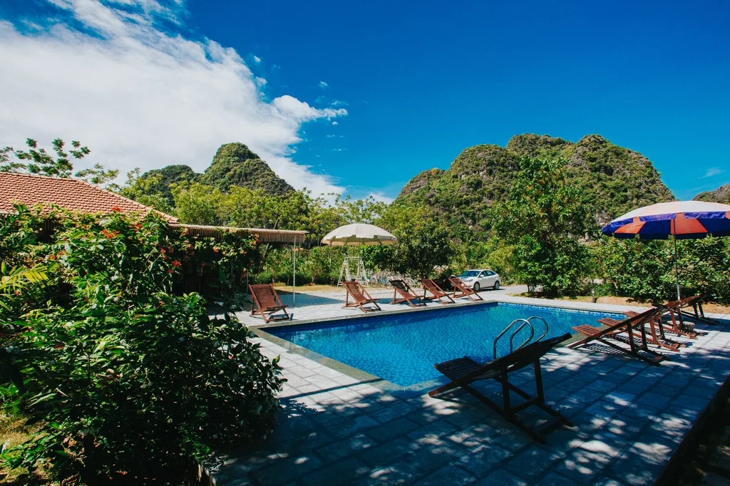 Bể bơi ngoài trời giữa vườn cây, ghế tắm nắng và dãy núi đá vôi phía sau tại Trang An Retreat