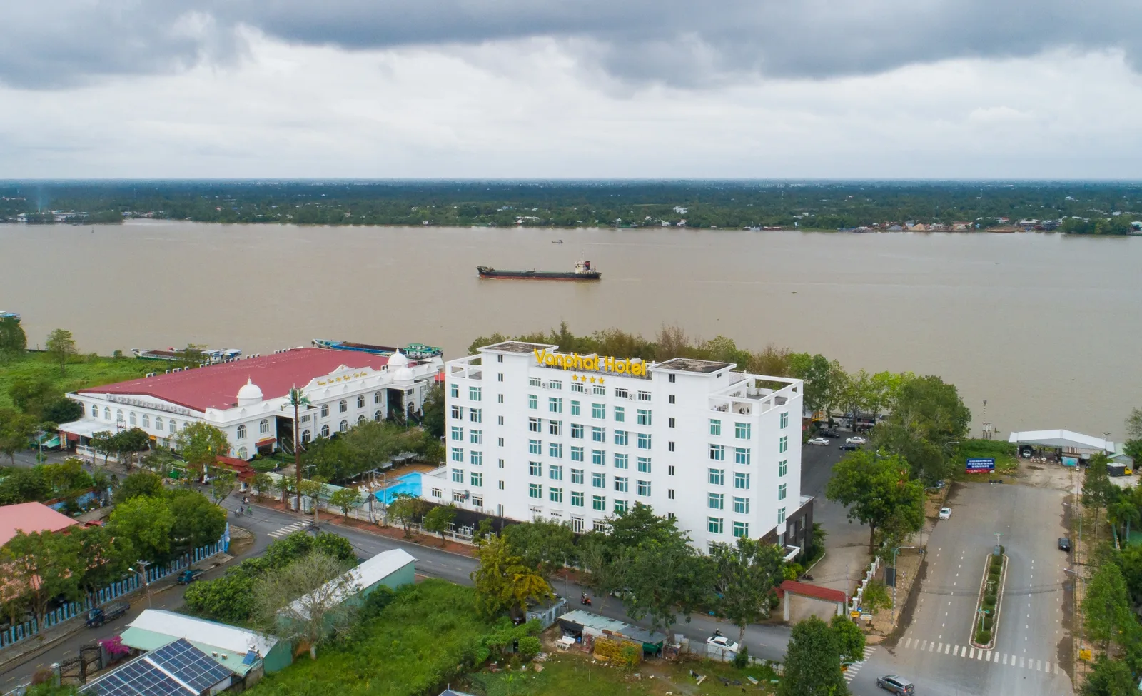 Toàn cảnh Vạn Phát Riverside Hotel nhìn từ trên cao, tòa nhà trắng cạnh bể bơi và sảnh tiệc mái đỏ, sông Hậu rộng phía sau