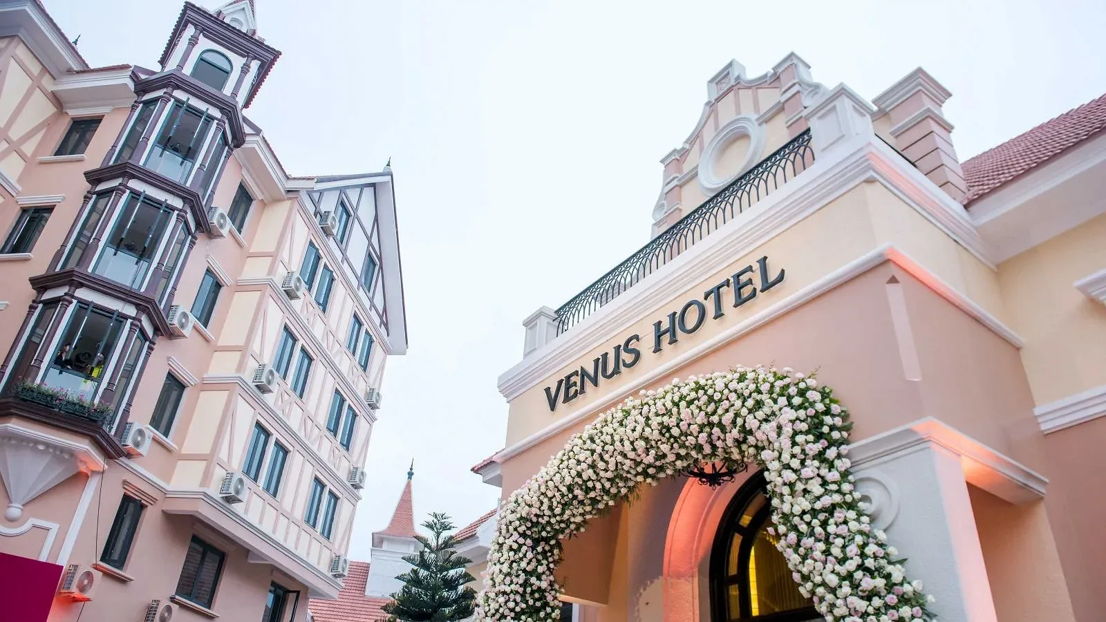 Mặt tiền Venus Hotel Tam Đảo với tòa nhà tân cổ điển màu hồng nhạt, tháp nhọn và cổng hoa trắng ở lối vào
