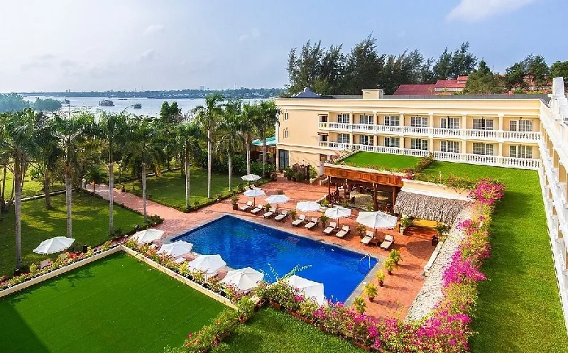 Hồ bơi ngoài trời giữa bãi cỏ và hàng cọ của Victoria Cần Thơ Resort, dãy nhà màu vàng bên phải, sông Hậu phía xa