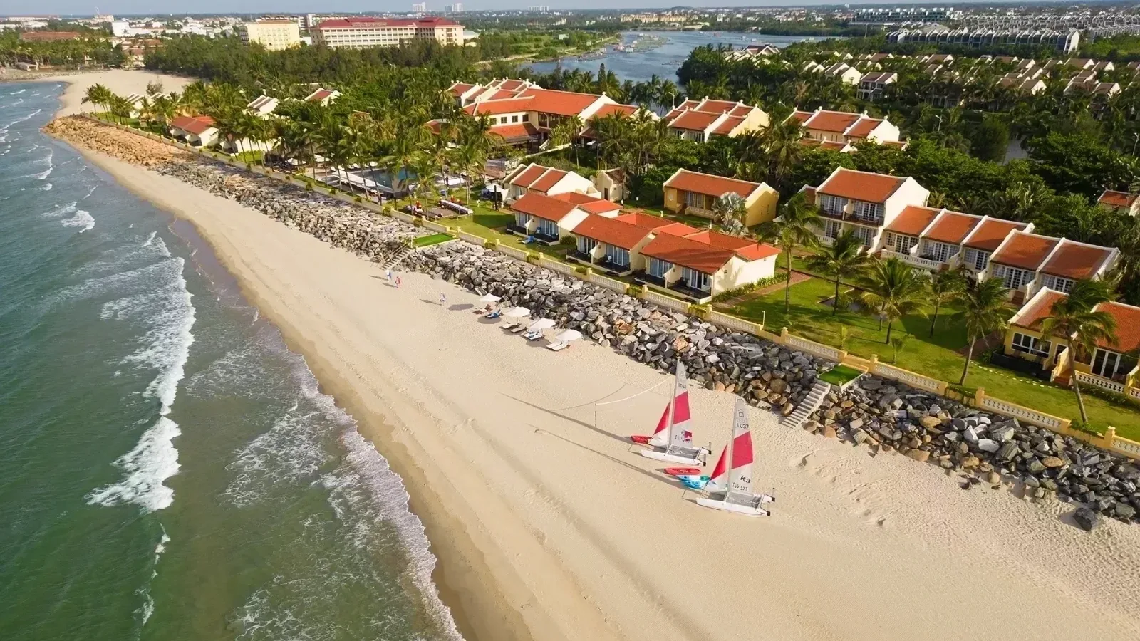 Ảnh flycam bãi cát Cửa Đại trước Victoria Hoi An Beach Resort & Spa, dãy nhà mái ngói đỏ giữa hàng dừa, hai thuyền buồm trắng đỏ trên cát