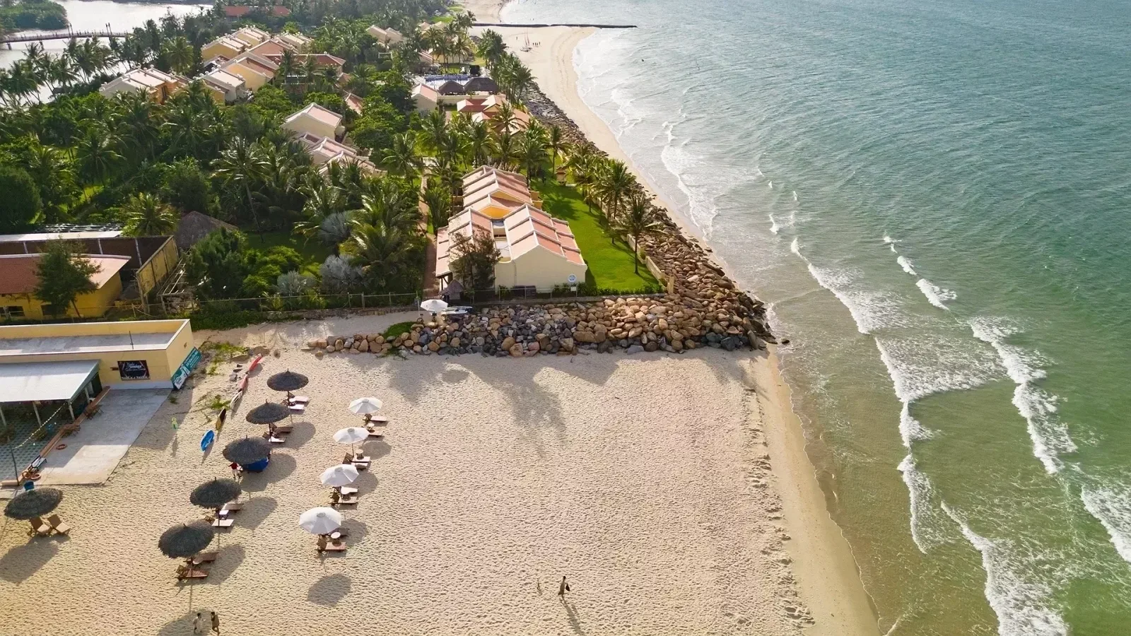 Toàn cảnh từ trên cao Victoria Hoi An Beach Resort & Spa: dãy nhà mái ngói giữa hàng dừa, kè đá, bãi cát có dù và ghế nằm cạnh biển