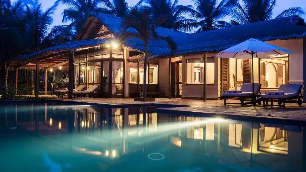 Villa mái tranh của Victoria Phan Thiet lên đèn buổi tối, hồ bơi phía trước và ghế nằm dưới ô trắng