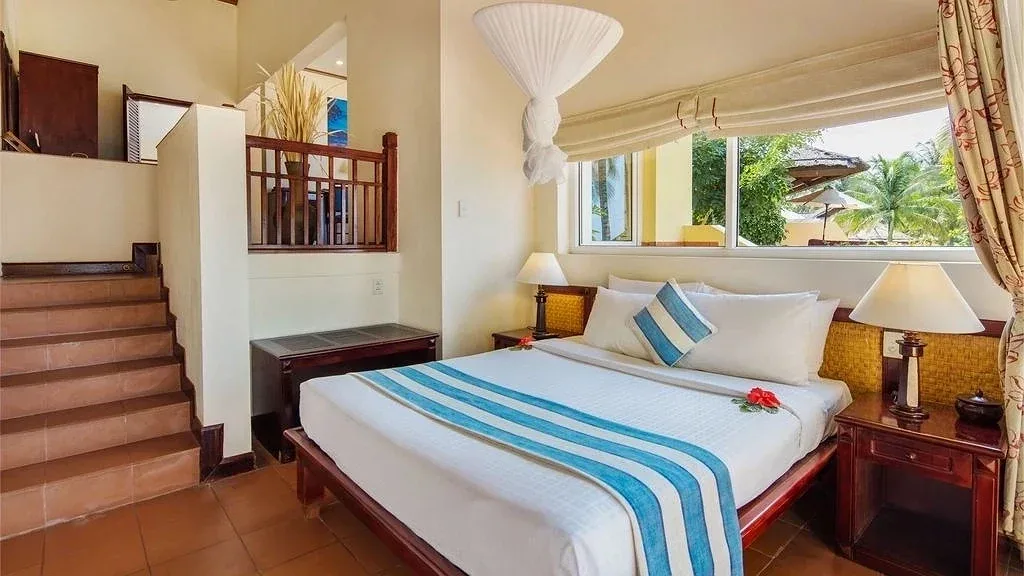 Phòng ngủ Family Bungalow tại Victoria Phan Thiet với giường lớn phủ khăn sọc xanh, màn chụp trắng và cầu thang gỗ dẫn lên gác lửng