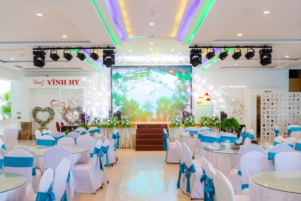 Sảnh tiệc trong nhà của Resort Vĩnh Hy, bàn tròn phủ khăn trắng và nơ xanh, sân khấu có màn hình lớn