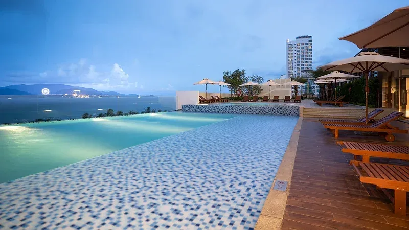 Hồ bơi vô cực trên cao của Vinpearl Beachfront Nha Trang lúc chiều tối, hàng ghế gỗ và ô dù bên cạnh, xa xa là vịnh và đảo lên đèn