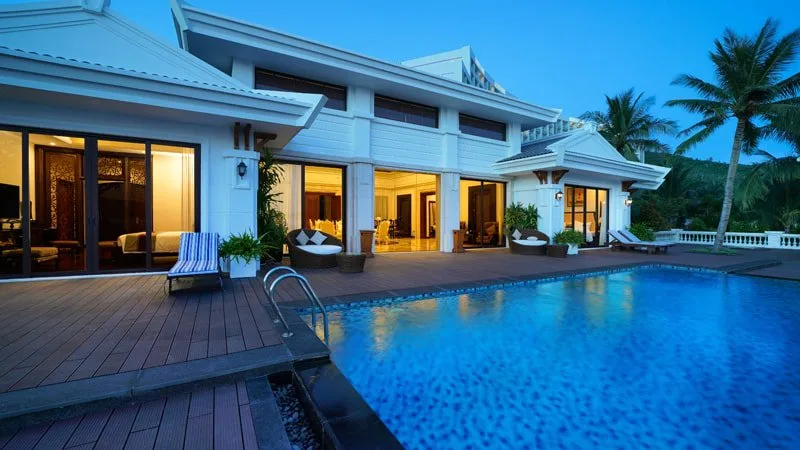 Biệt thự tường trắng tại Vinpearl Golf Land Resort & Villas Nha Trang lúc chạng vạng, sàn gỗ quanh bể bơi riêng xanh, cửa kính nhìn vào phòng ngủ và phòng khách