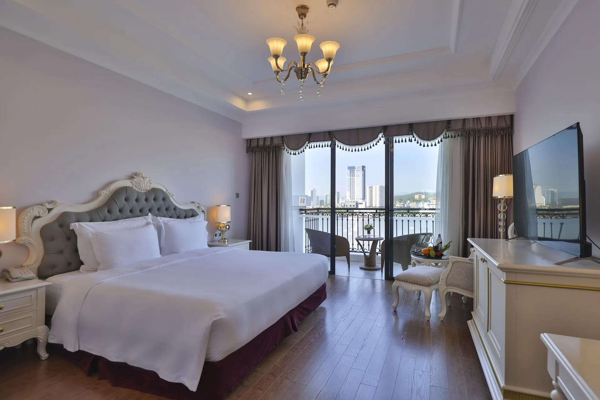 Phòng Deluxe Hướng Biển Giường Đôi với giường lớn đầu giường bọc nỉ xám, cửa kính mở ra ban công có bàn ghế mây nhìn sang phố Bãi Cháy