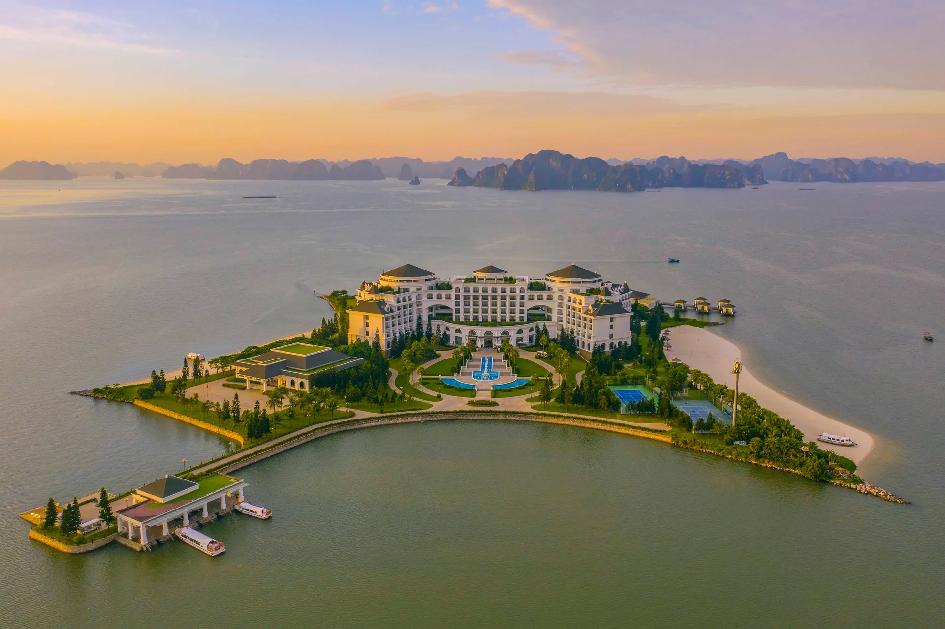 Toàn cảnh đảo Rều lúc hoàng hôn với tòa nhà tân cổ điển, bãi cát trắng, bến cano và dãy núi đá vịnh Hạ Long phía xa