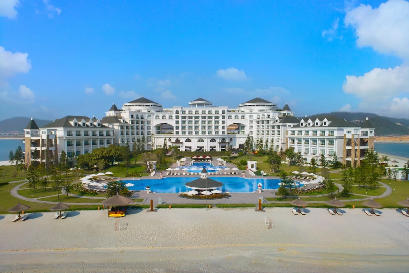 Tòa nhà tân cổ điển màu trắng của Vinpearl Resort & Spa Hạ Long nhìn từ bãi cát, bể bơi ngoài trời lớn có chòi giữa bể, ô dù và ghế nằm