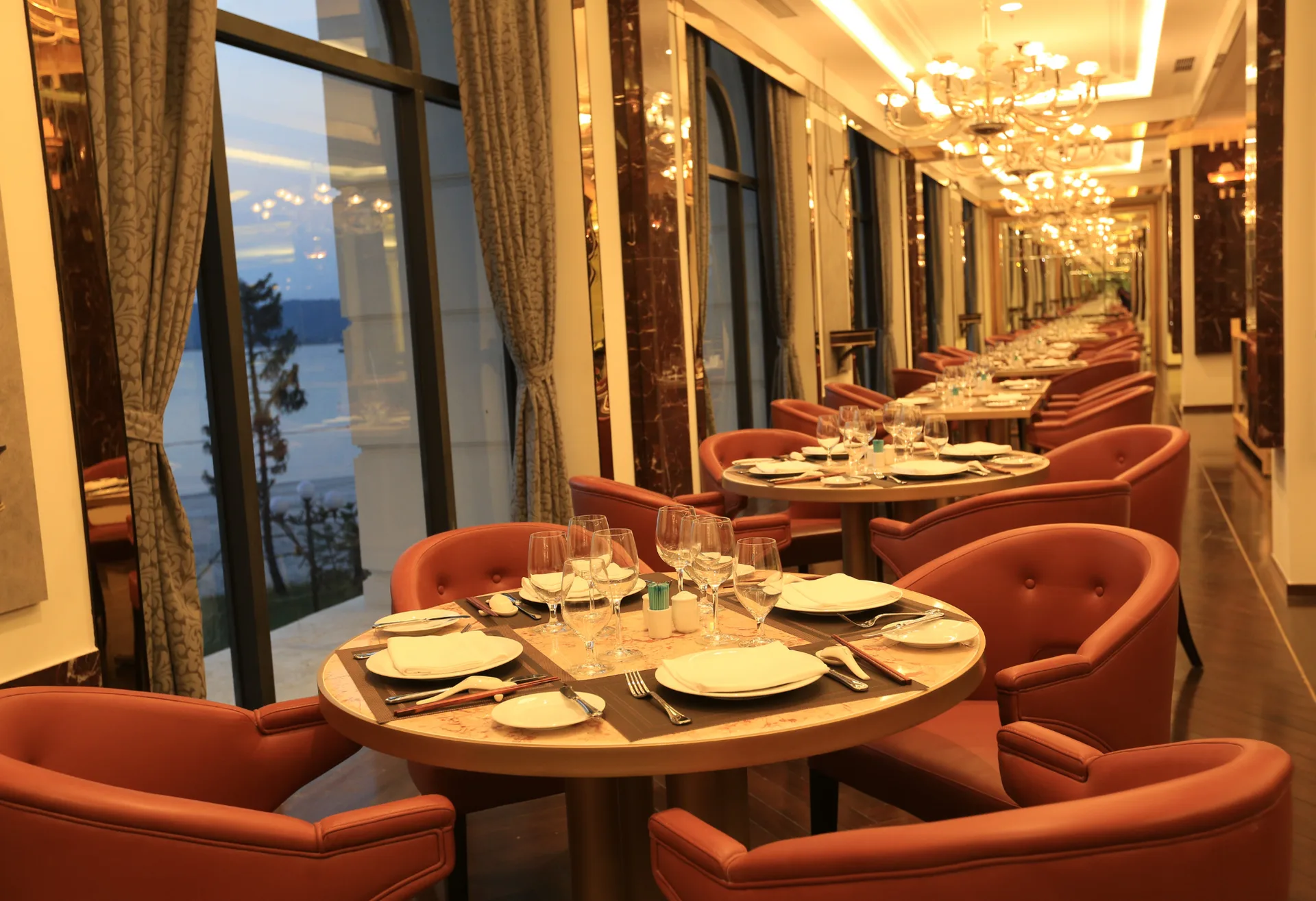 Dãy bàn tròn ghế da màu cam, đèn chùm pha lê và cửa kính lớn nhìn ra mặt nước lúc chiều tối ở nhà hàng Bayview