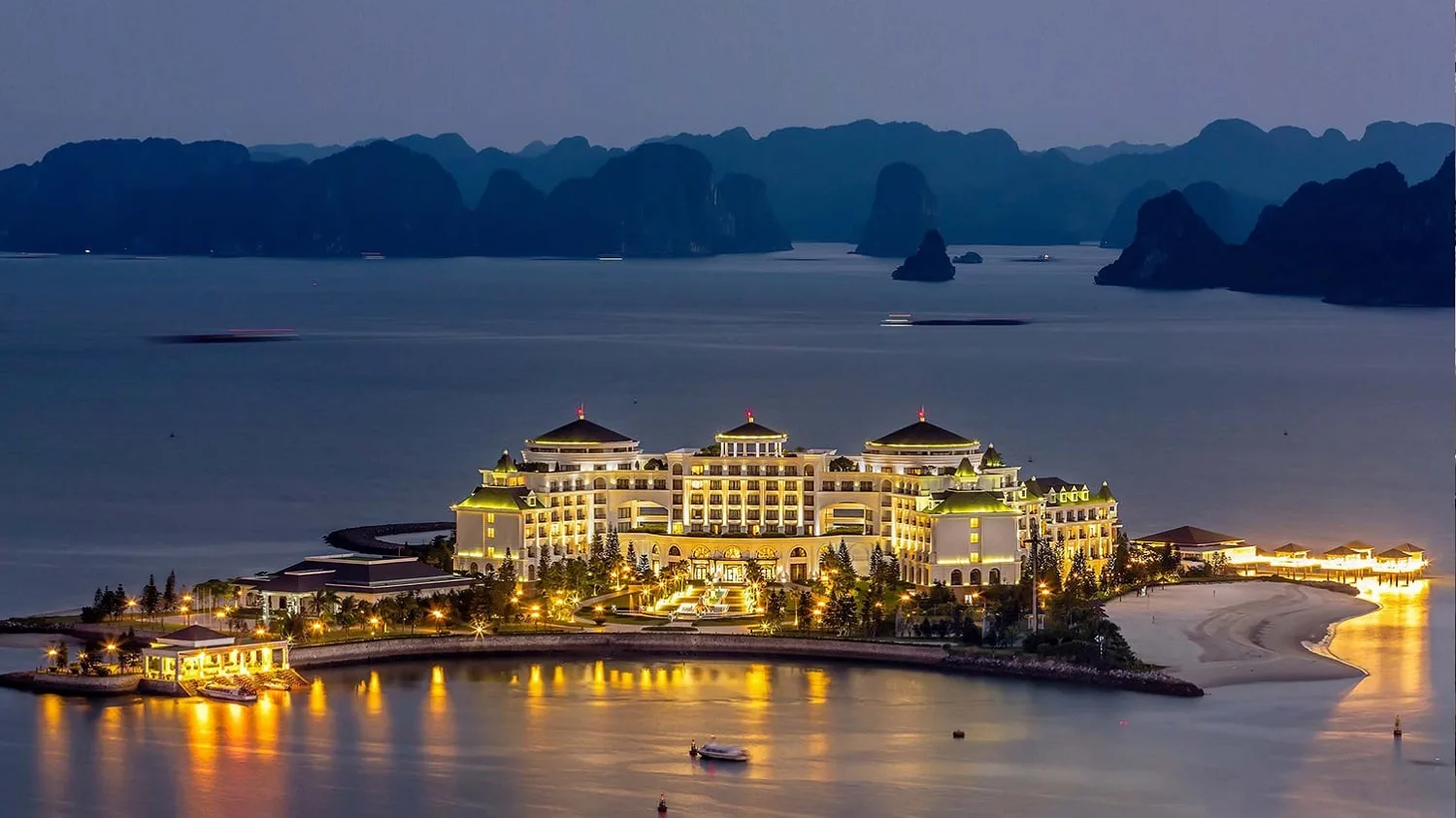 Vinpearl Resort & Spa Hạ Long trên đảo Rều lên đèn về đêm, bến cano và dãy nhà chòi trên biển, phía sau là núi đá vịnh Hạ Long