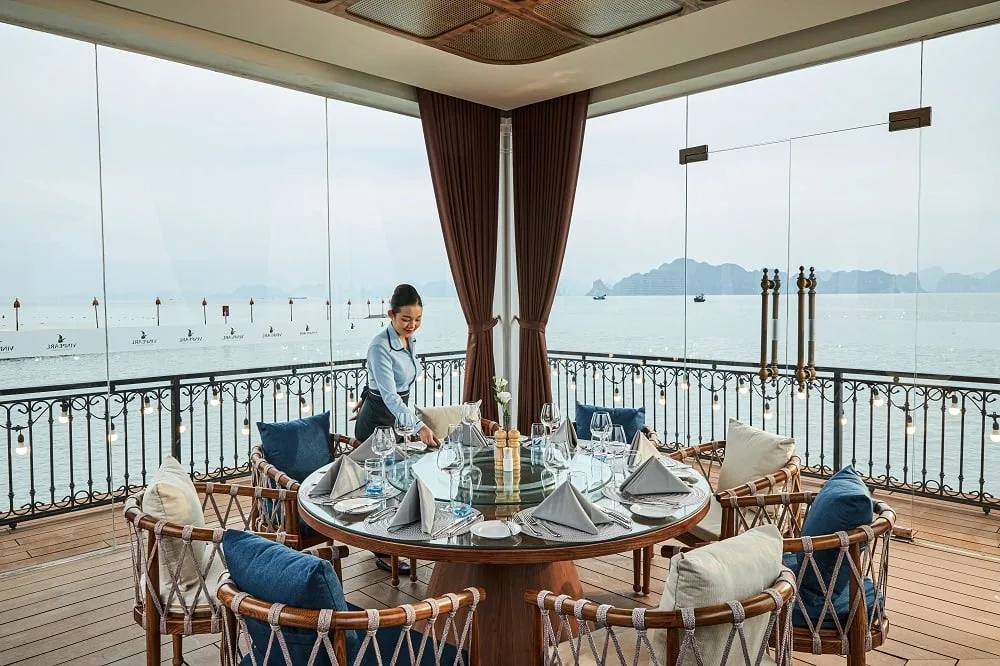Bàn ăn tròn trong nhà chòi Pavilion vách kính trên mặt biển, nhìn ra vịnh có núi đá và tàu thuyền