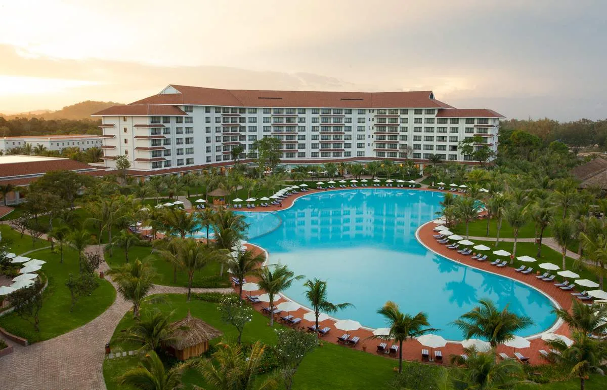 Bể bơi ngoài trời lớn của Vinpearl Resort & Spa Phú Quốc uốn lượn giữa bãi cỏ và hàng dừa, phía sau là tòa khách sạn mái ngói đỏ