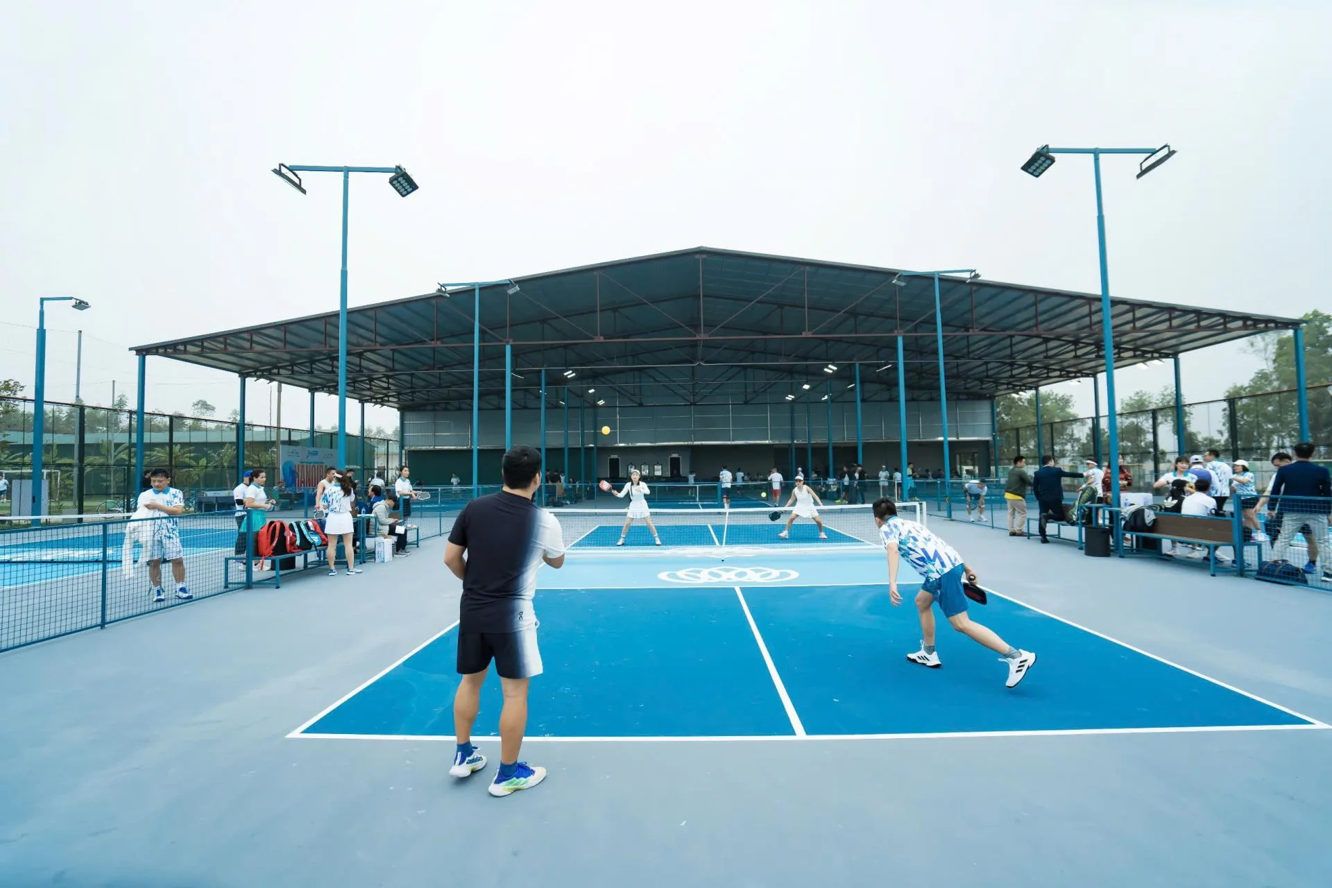 Sân pickleball nền xanh dưới mái tôn khung thép lớn, có cột đèn, khách đang thi đấu ở Vườn Vua