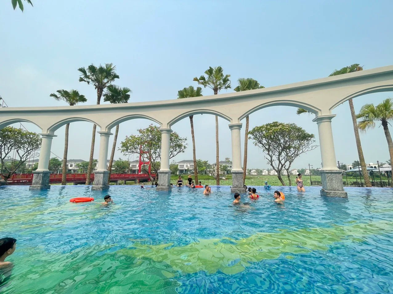 Bể bơi ngoài trời có hàng vòm cột trắng chạy qua lòng bể, khách đang bơi, cầu đỏ và đầm sen phía sau ở Vườn Vua