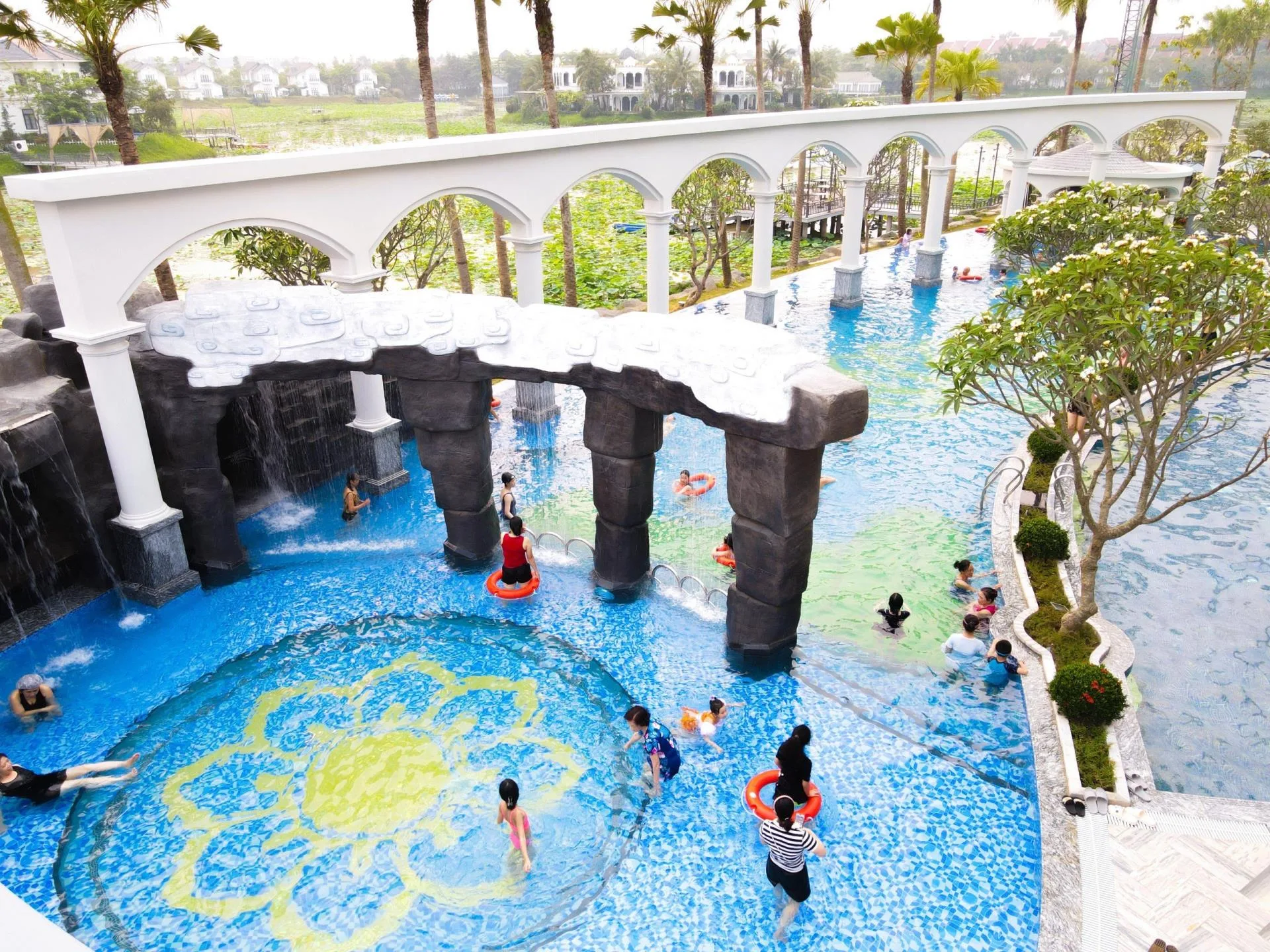 Bể Golden Palm Onsen ở Vườn Vua nhìn từ trên cao: hàng vòm trắng, trụ đá giả có thác nước, đáy bể khảm hoa sen vàng, nhiều khách đang tắm