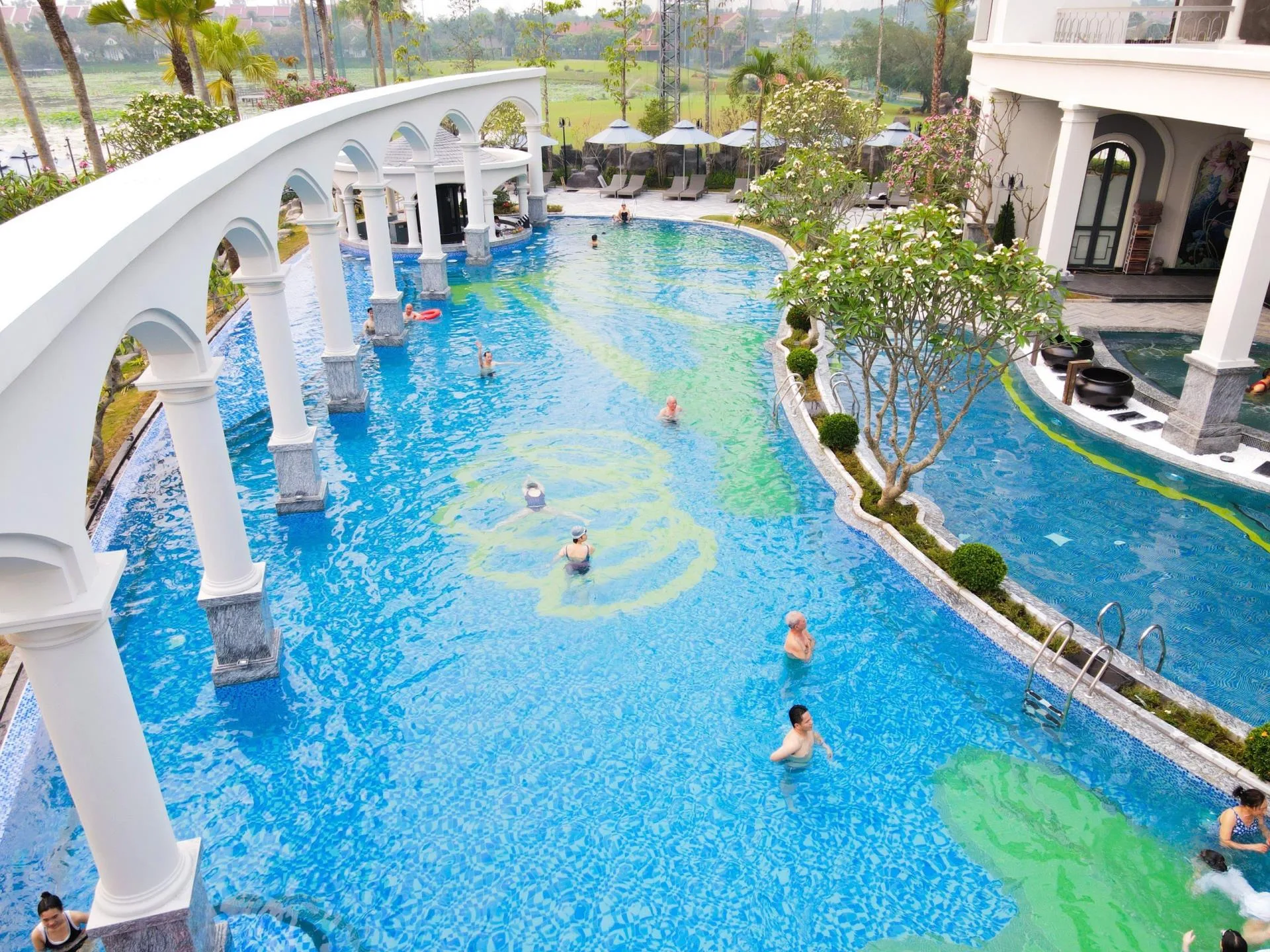 The Golden Palm Onsen Swimming Pool tại Vườn Vua Resort & Villas với hàng vòm cột trắng chạy qua lòng bể, khách đang bơi