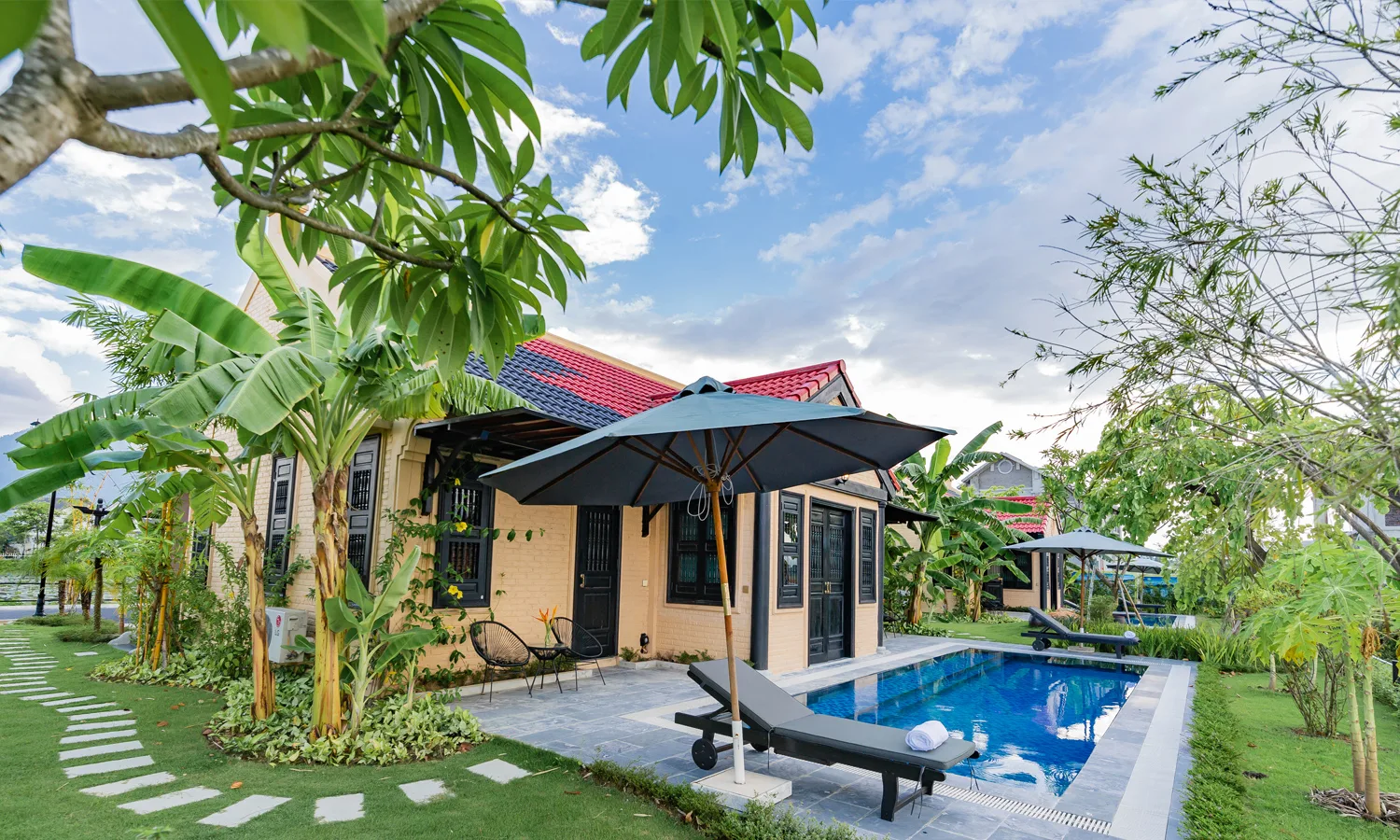 Biệt thự Hồng Liên 1 tầng tường vàng mái ngói đỏ với bể bơi riêng, ghế tắm nắng và dù trong vườn chuối