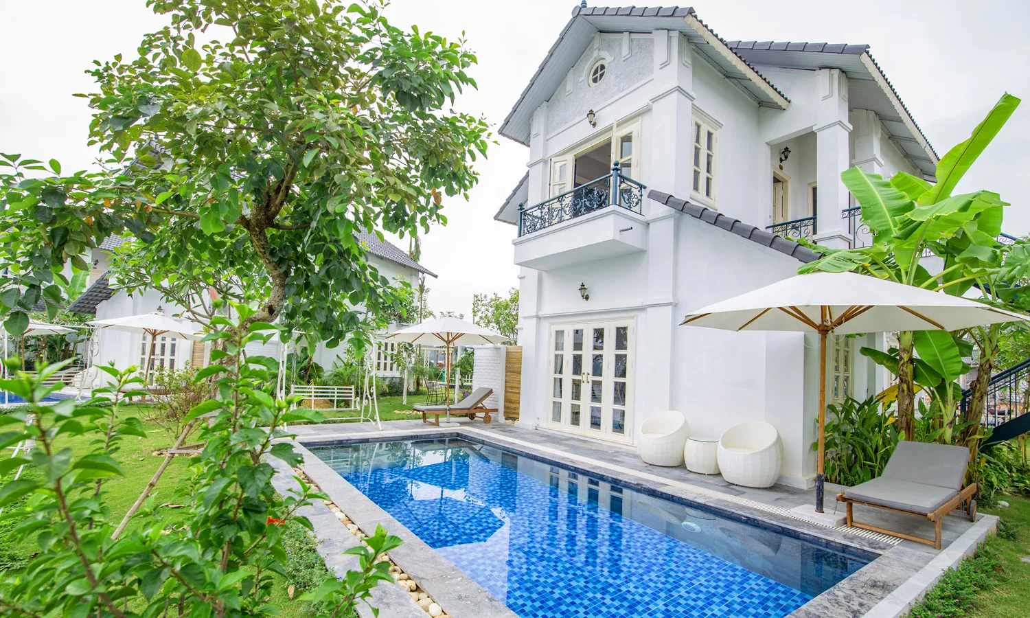 Biệt thự Thanh Liên trắng 2 tầng ban công sắt uốn, bể bơi riêng lát gạch xanh, ghế mây và dù trắng