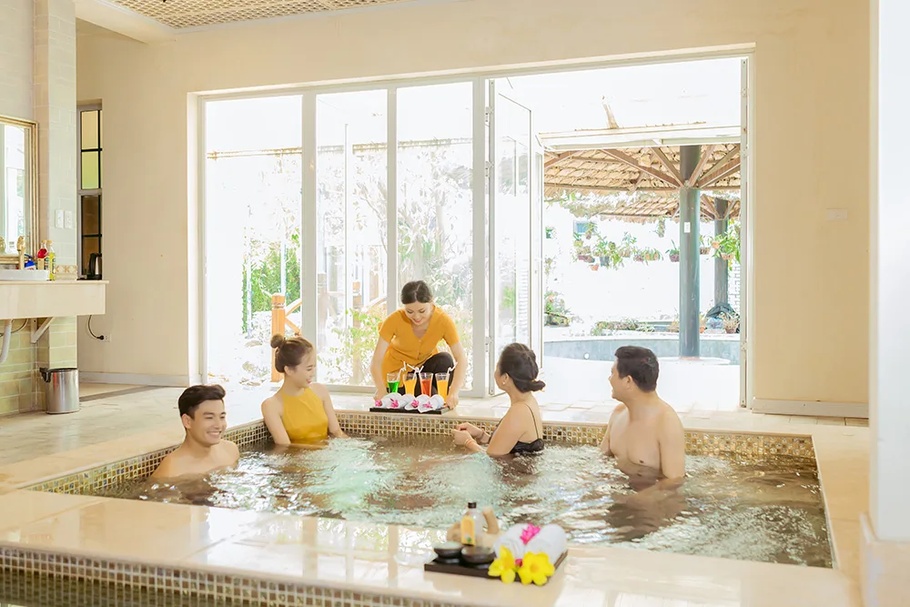 Bể sục khoáng nóng trong nhà ở Vườn Vua, bốn khách ngâm mình, nhân viên mang khay nước trái cây, cửa kính mở ra sân vườn