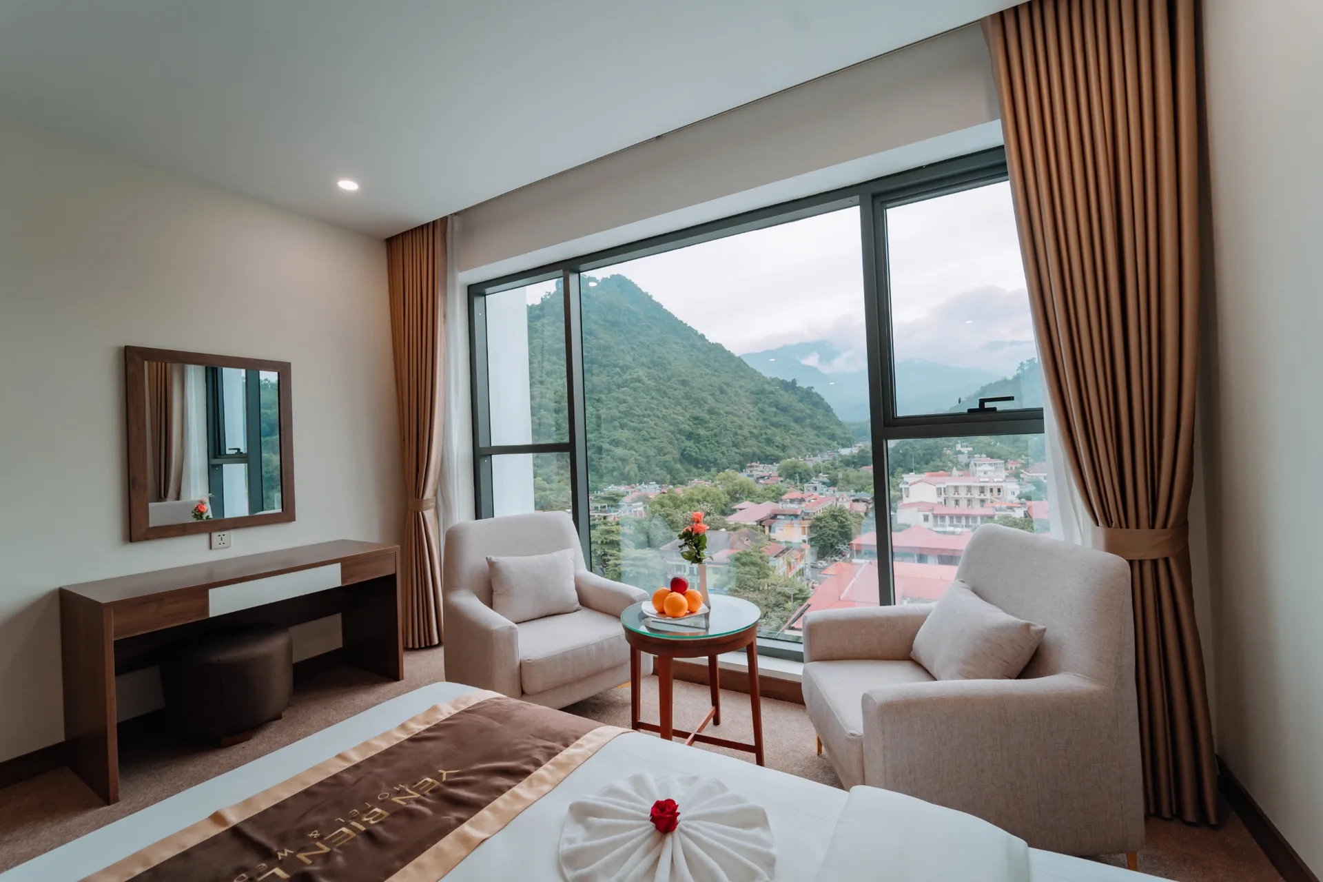 Phòng nghỉ Yên Biên Luxury Hotel có cửa kính lớn nhìn ra núi xanh và mái nhà thành phố, hai ghế bành bên bàn trà